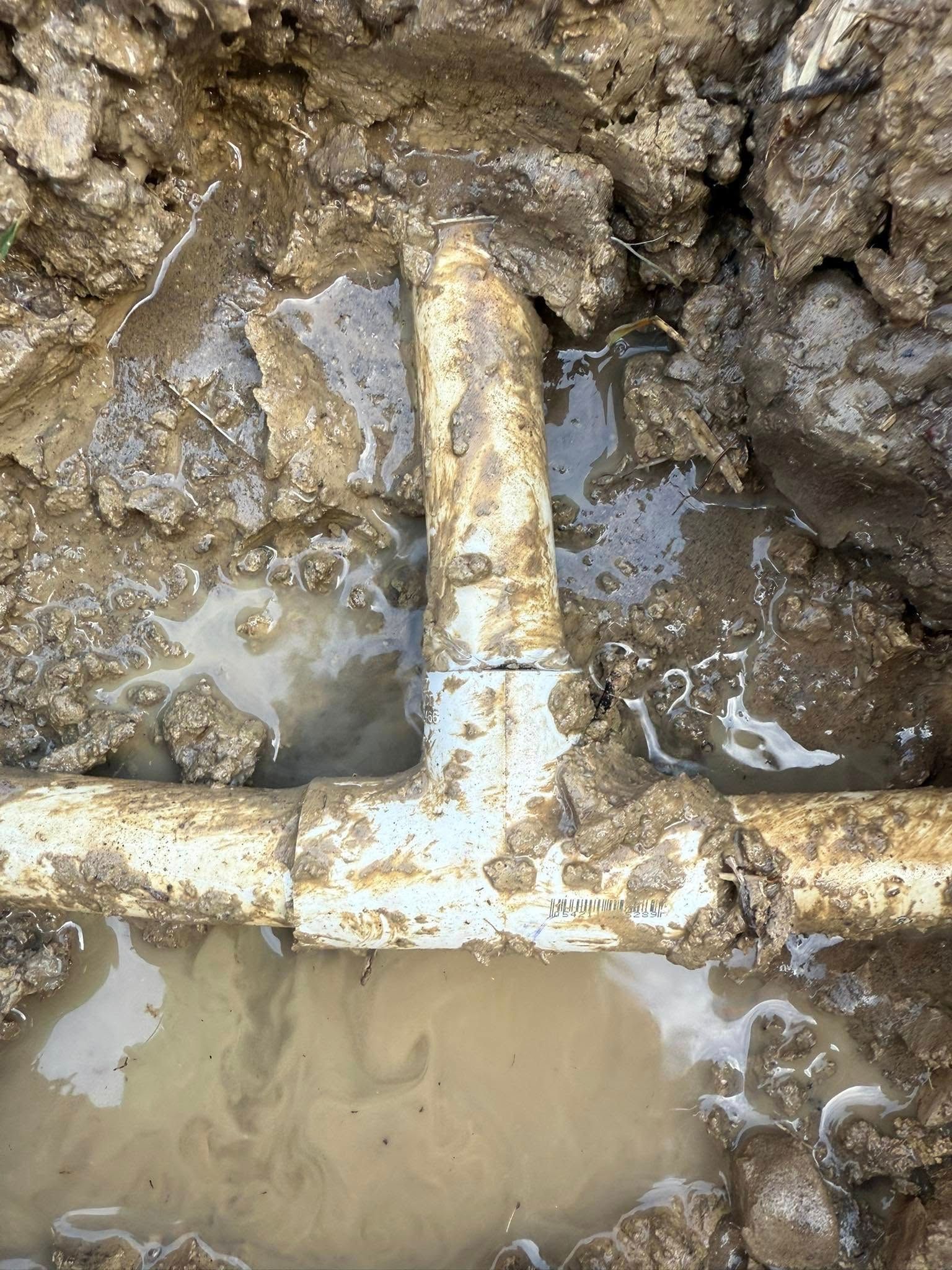 Damaged PVC pipe fitting in muddy ground, leaking water.