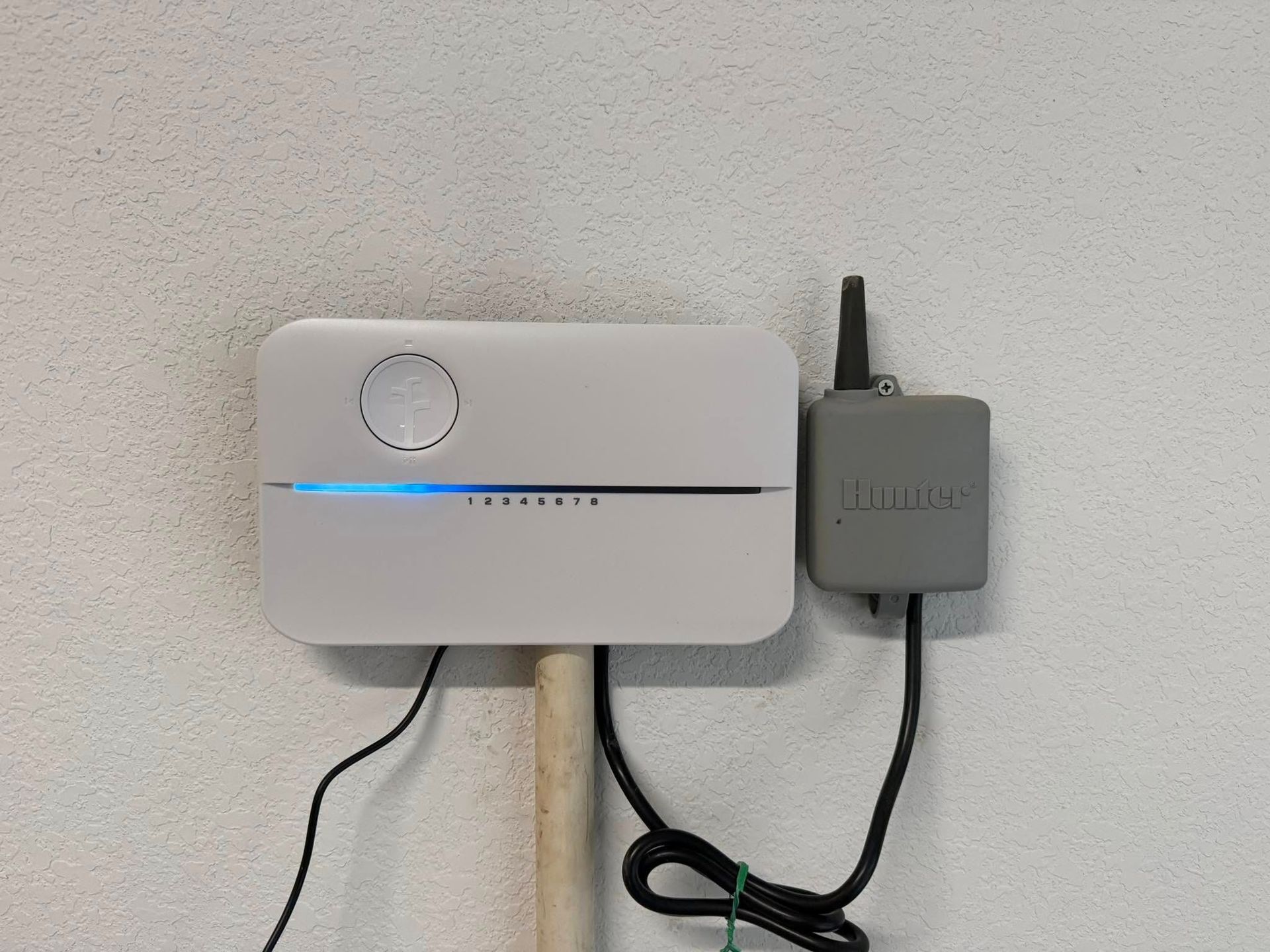 White Rachio sprinkler controller mounted on wall with a gray wireless sensor. A blue light is on the controller.