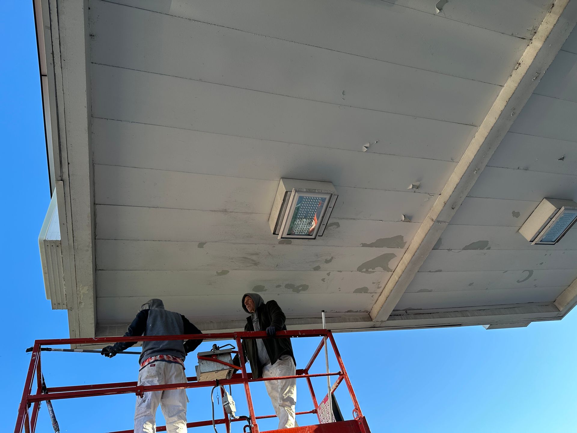 Two workers on a lift painting the underside of a white canopy against a blue sky.