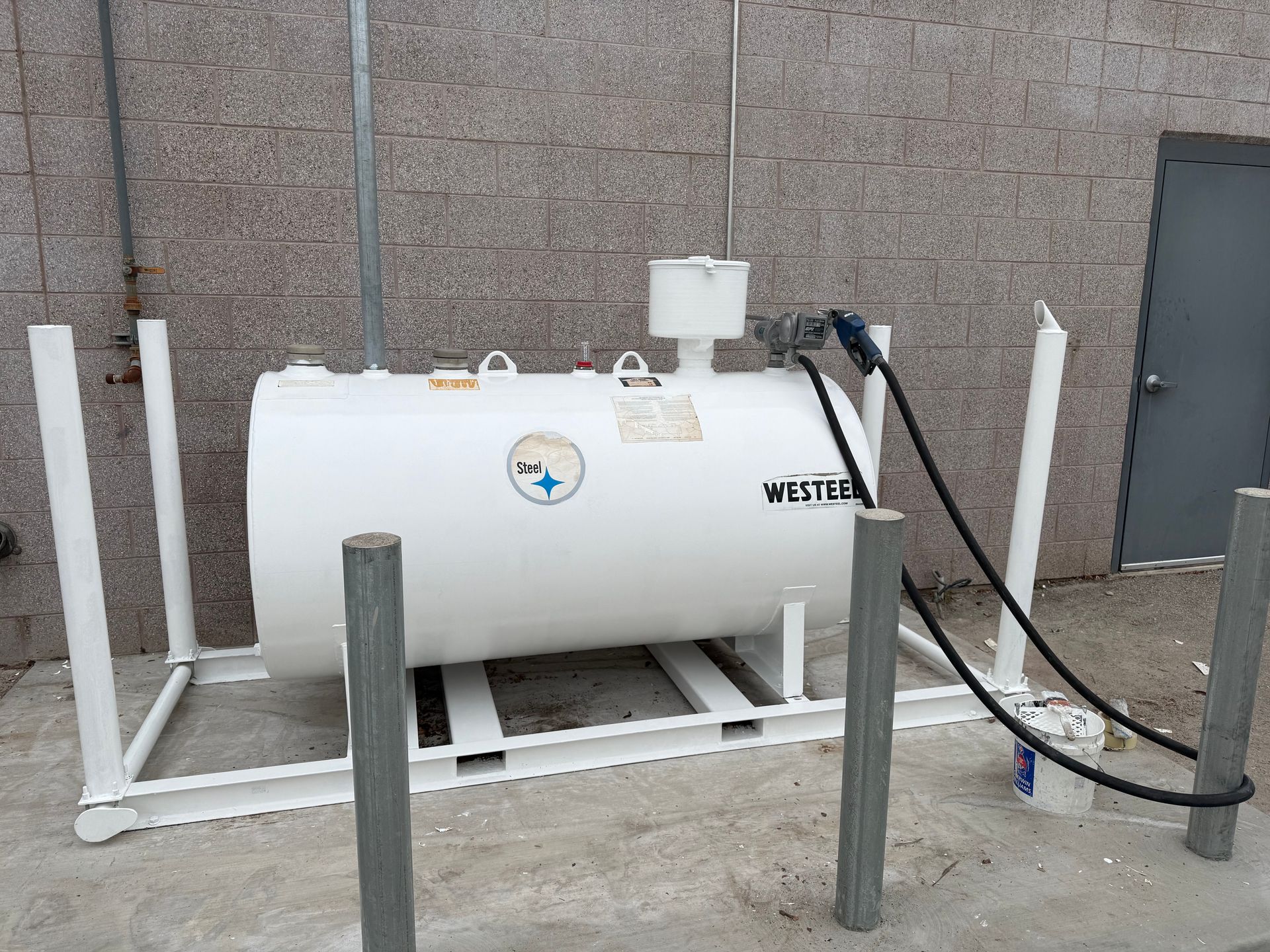 White fuel tank with nozzle and pump, on a white metal frame, next to a building.