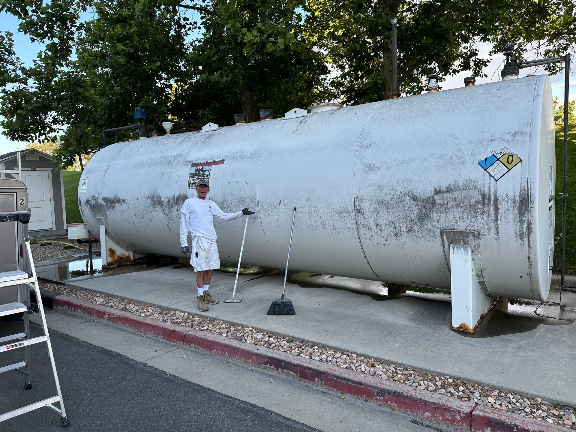 Person standing near large, white storage tank; brush and broom visible. Exterior shot.