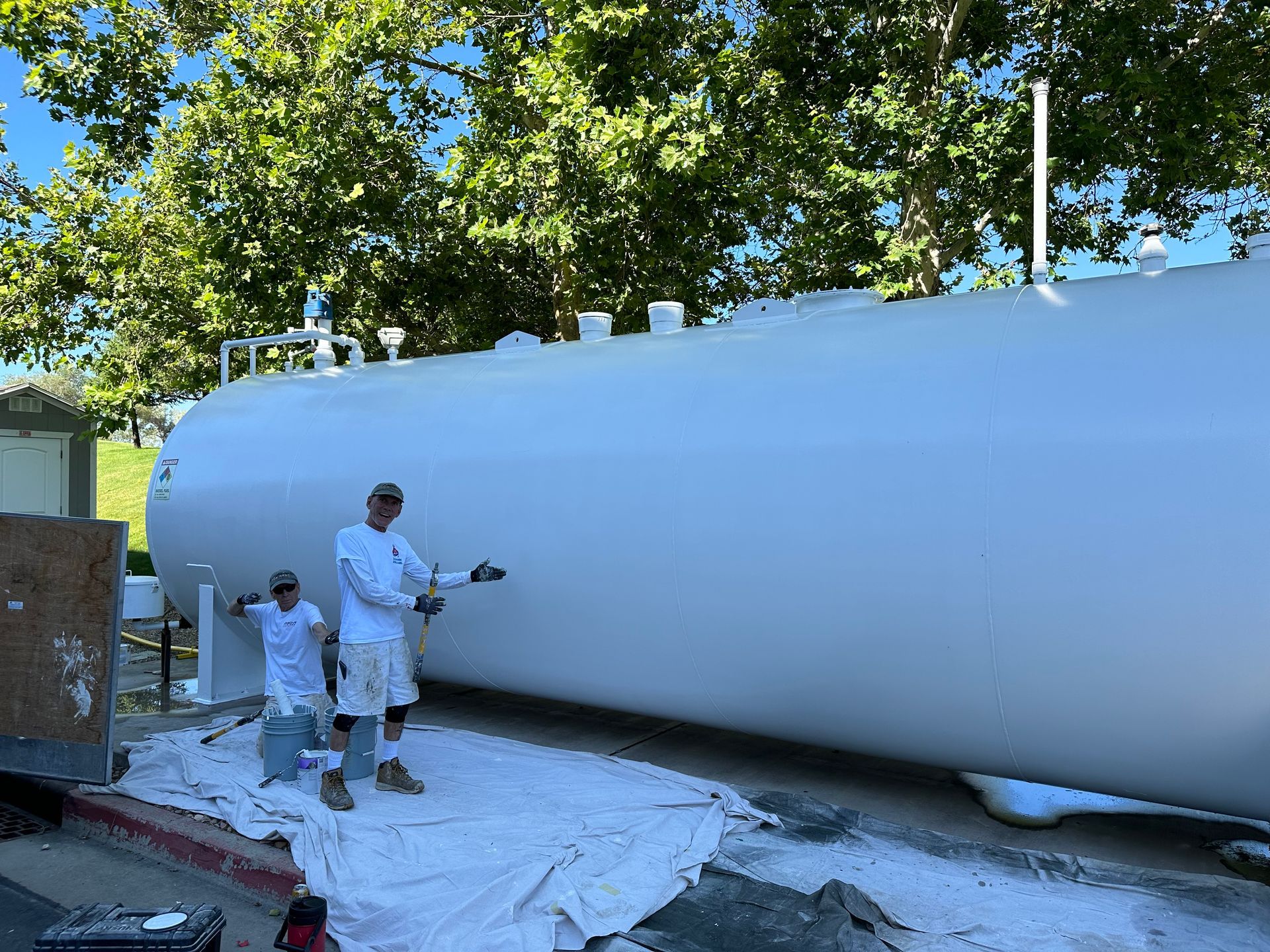Two people in white shirts and shorts next to a large, white tank, possibly after painting it.