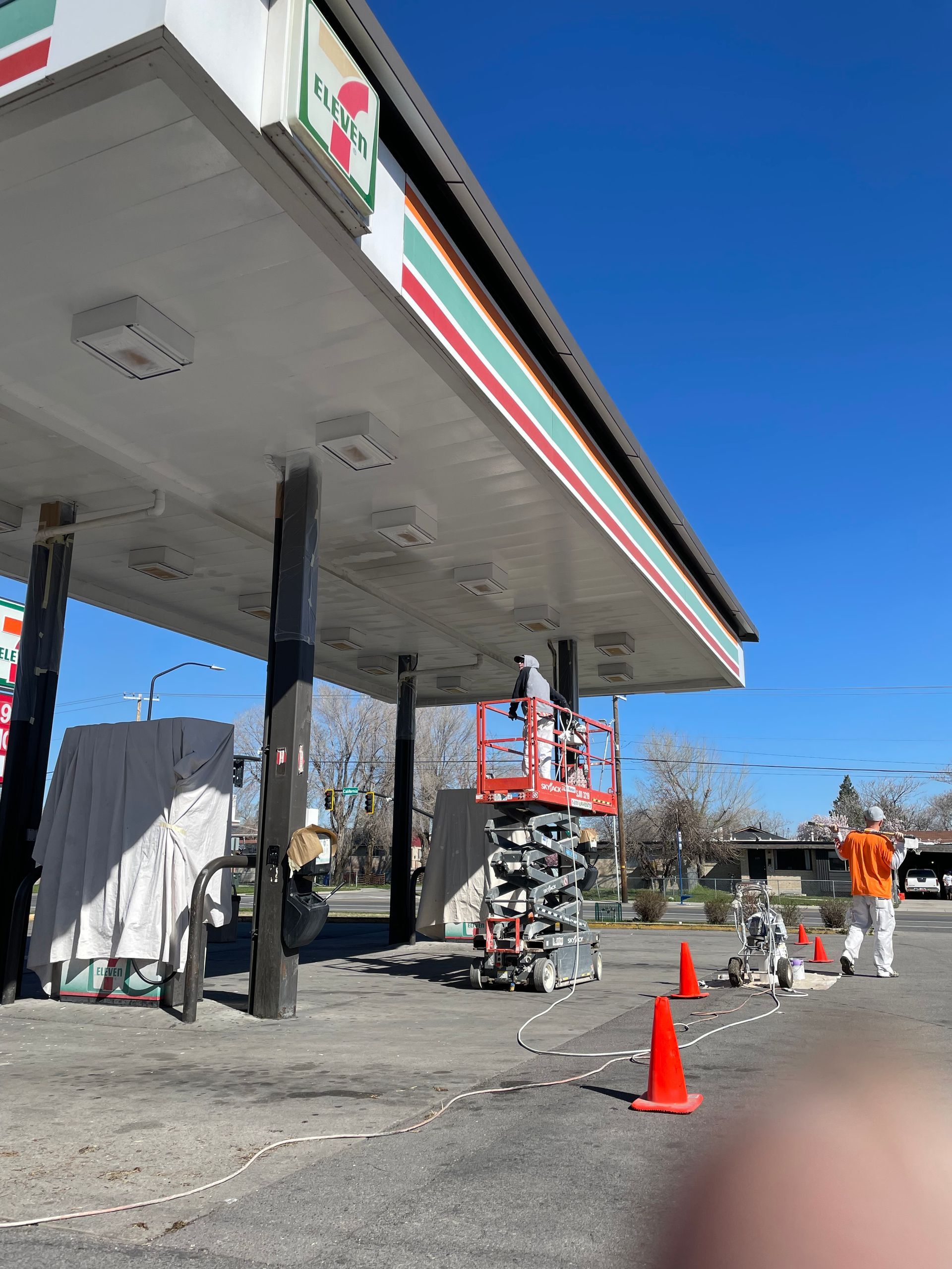 7-Eleven gas station canopy under construction; workers on lift, orange cones on ground.