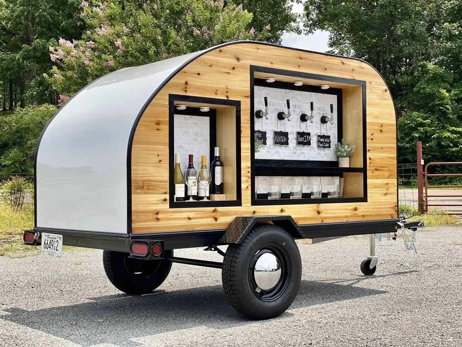 Wooden mobile bar on a trailer with wine bottles, taps, and a white curved roof.