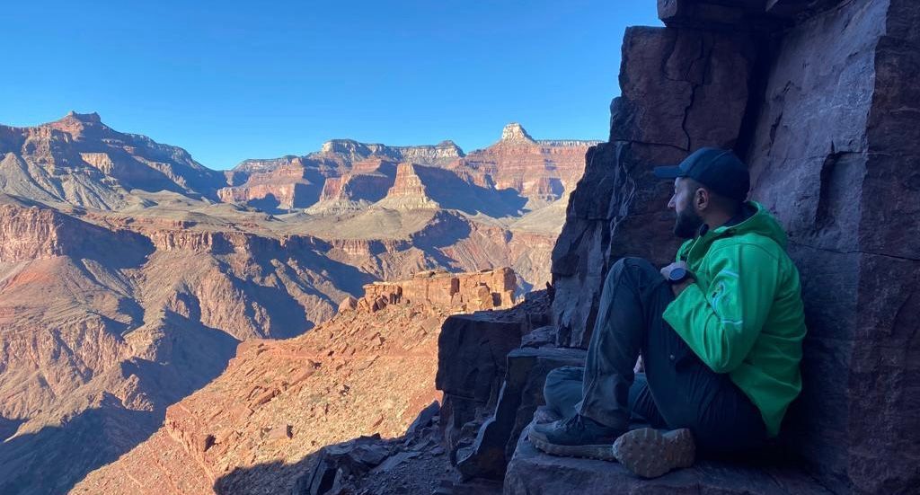 A man in a green jacket is sitting on the edge of a cliff overlooking a mountain range.