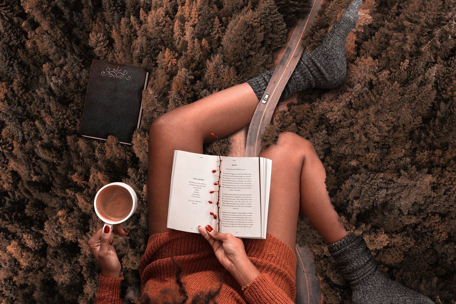 A woman is laying on the ground reading a book and drinking coffee.