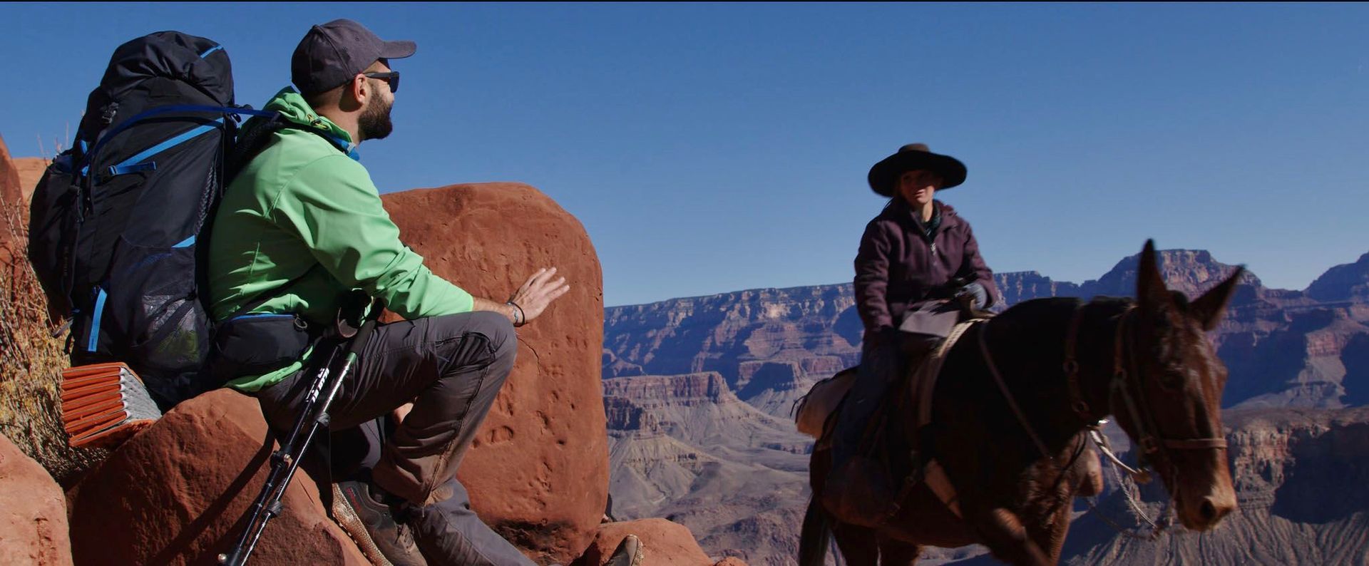 A man with a backpack is sitting on a rock next to a man riding a horse.