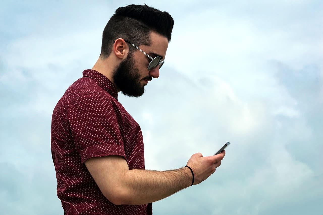 A man with a beard and sunglasses is looking at his cell phone.