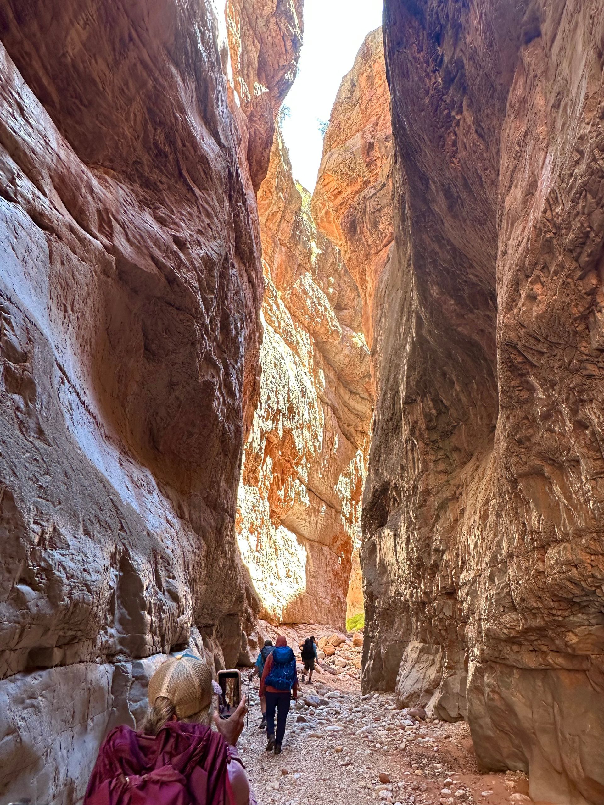 Hikers walking through a narrow, red-rock canyon. Sunlight streams from above.