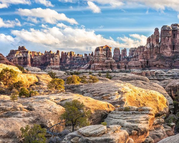 Sunlit red rock canyon with layered cliffs, scrubby foreground, and a blue sky with wispy clouds