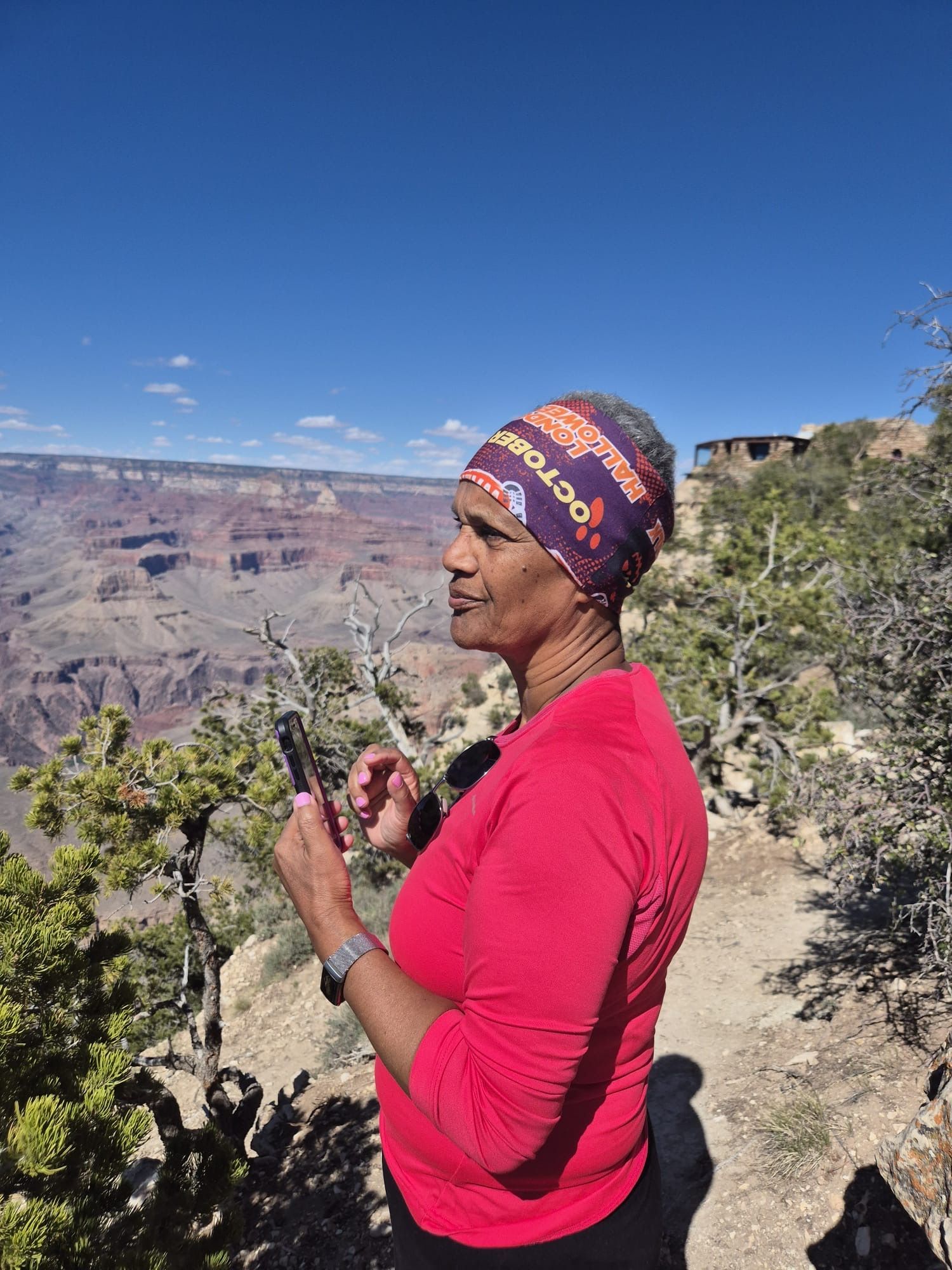 Woman with patterned head covering and red shirt at the Grand Canyon, looking at a phone. Blue sky.