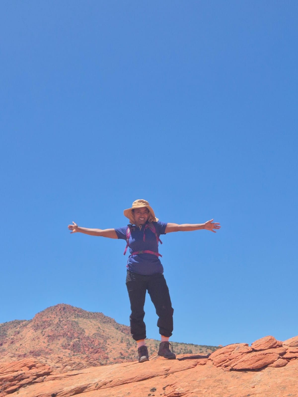 Person with arms outstretched, smiles on a red rock landscape under a bright blue sky.