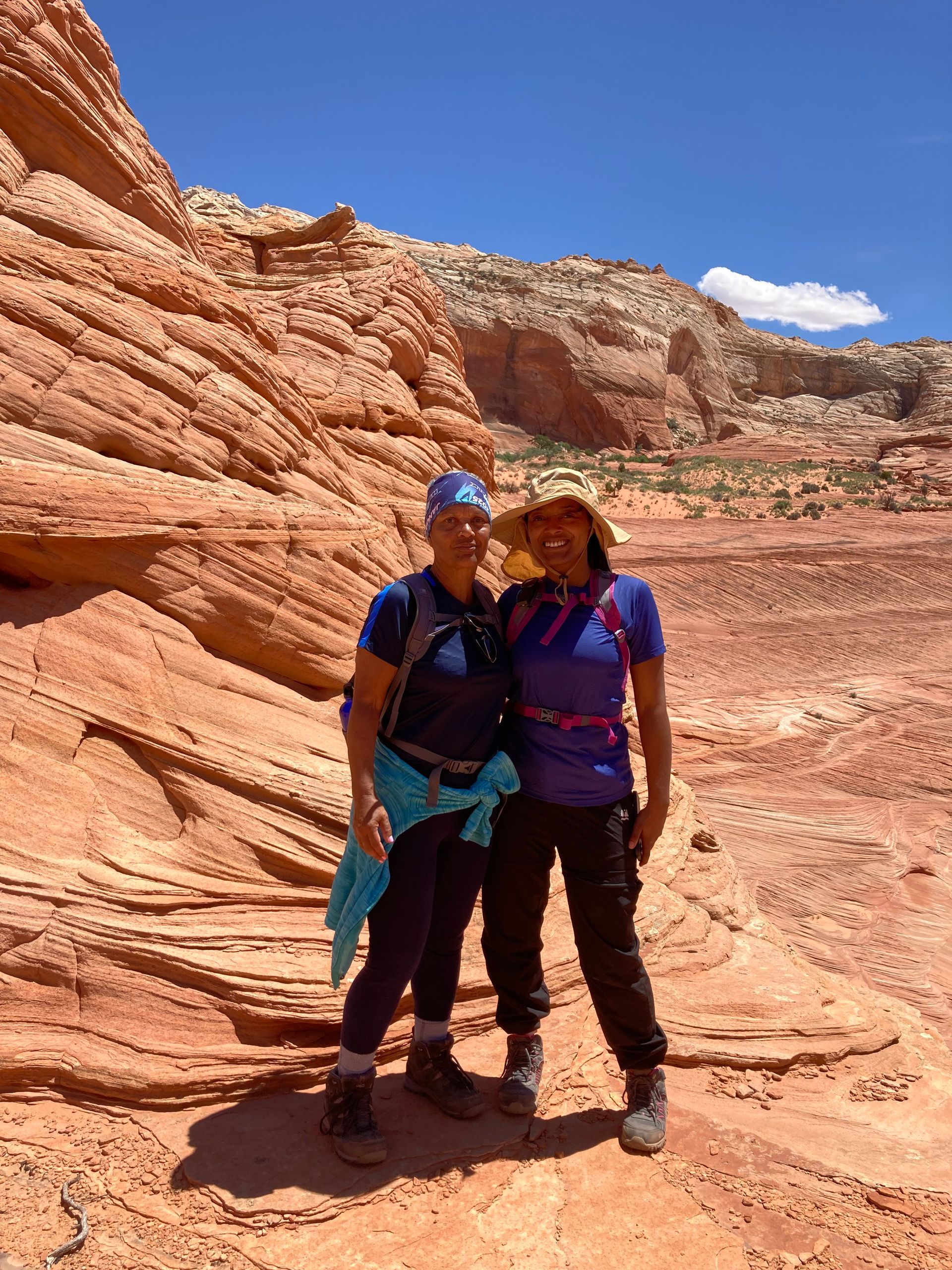 Two people stand posing against a red rock formation, sunny, desert environment.