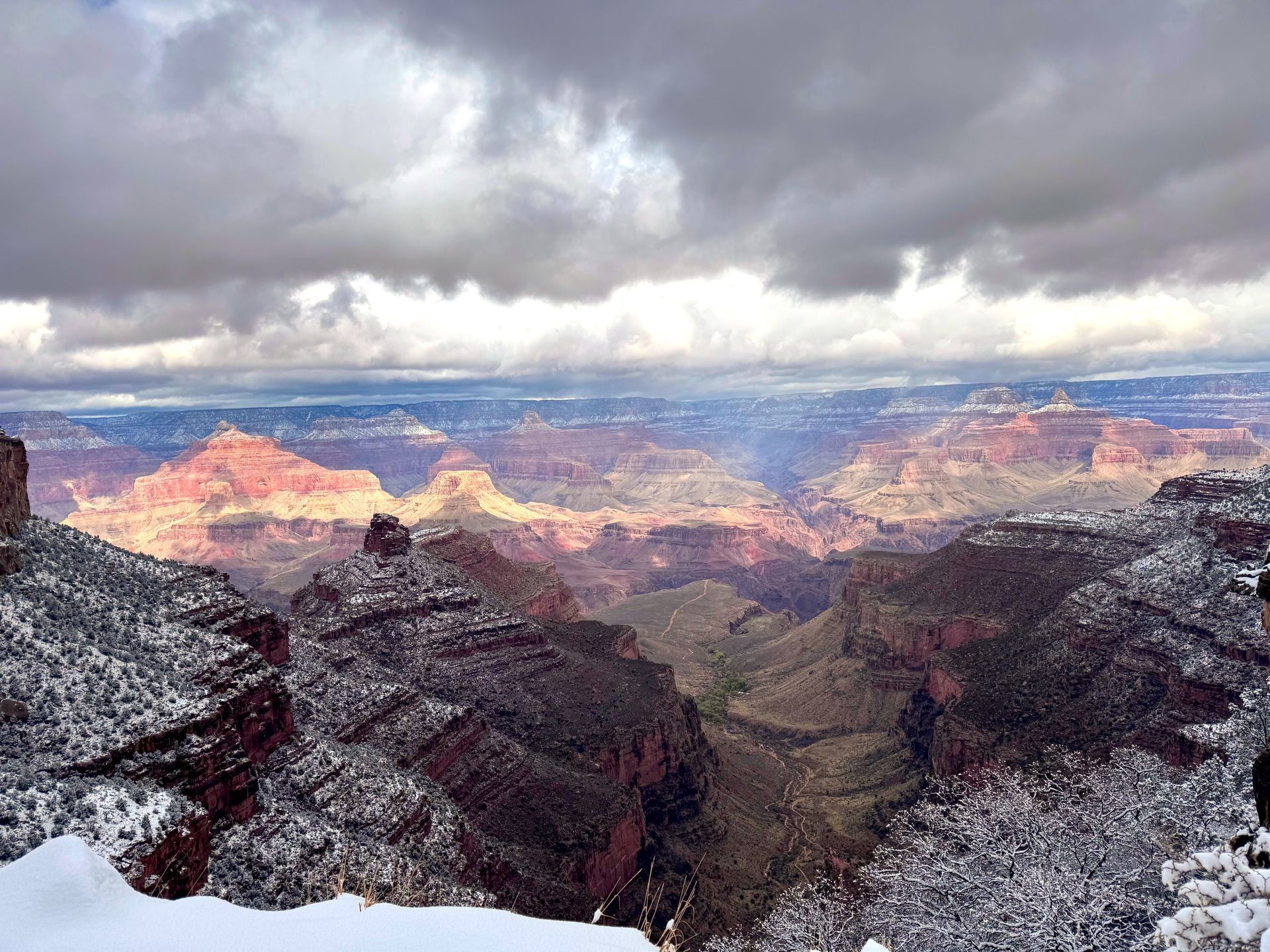 Snowy Grand Canyon vista, reddish rock formations, under cloudy, sunlit sky.