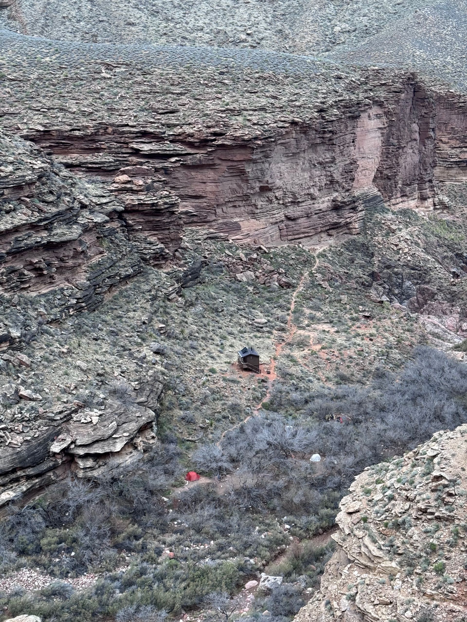 Rocky canyon landscape with layered rock formations, green vegetation, and a dark structure with a red flag.