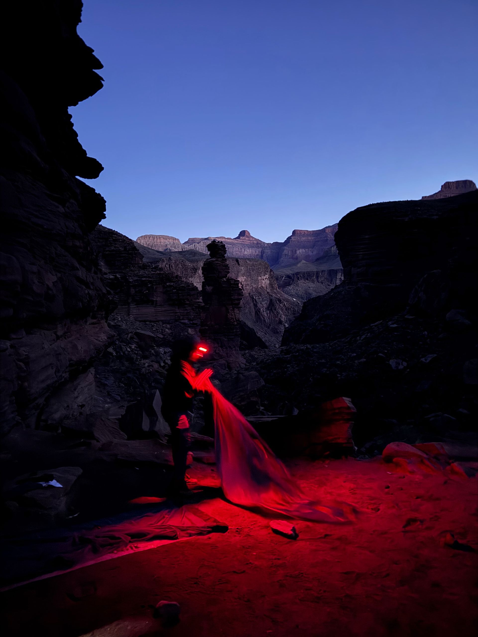 Person in dark canyon, illuminated by red light, pointing it towards a rock formation under a twilight sky.
