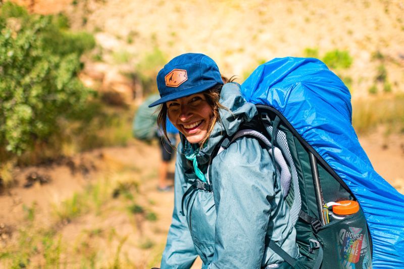 A woman wearing a blue hat and a backpack is smiling.
