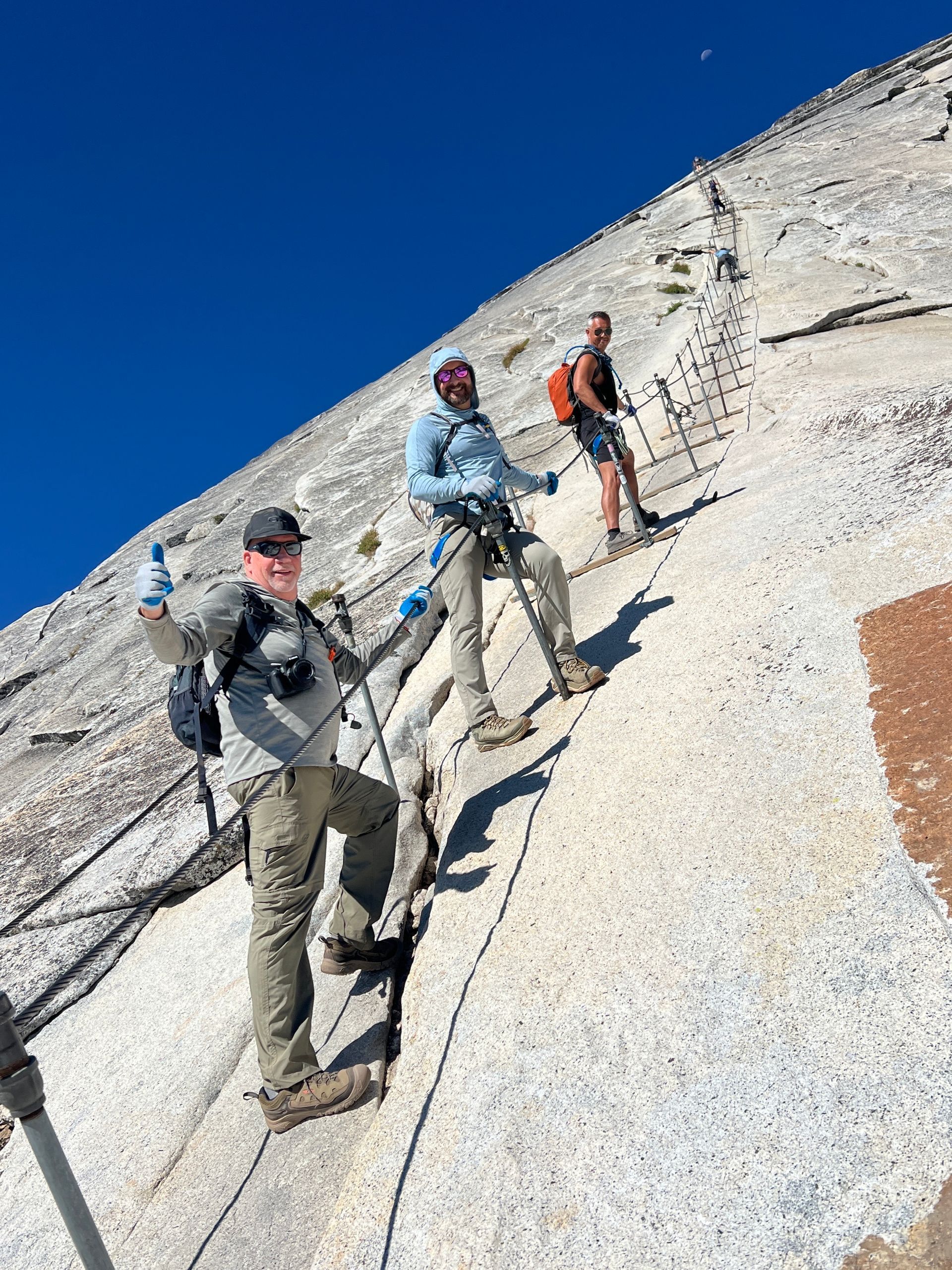 Three hikers ascending a steep granite slope using cables and posts, under a blue sky.