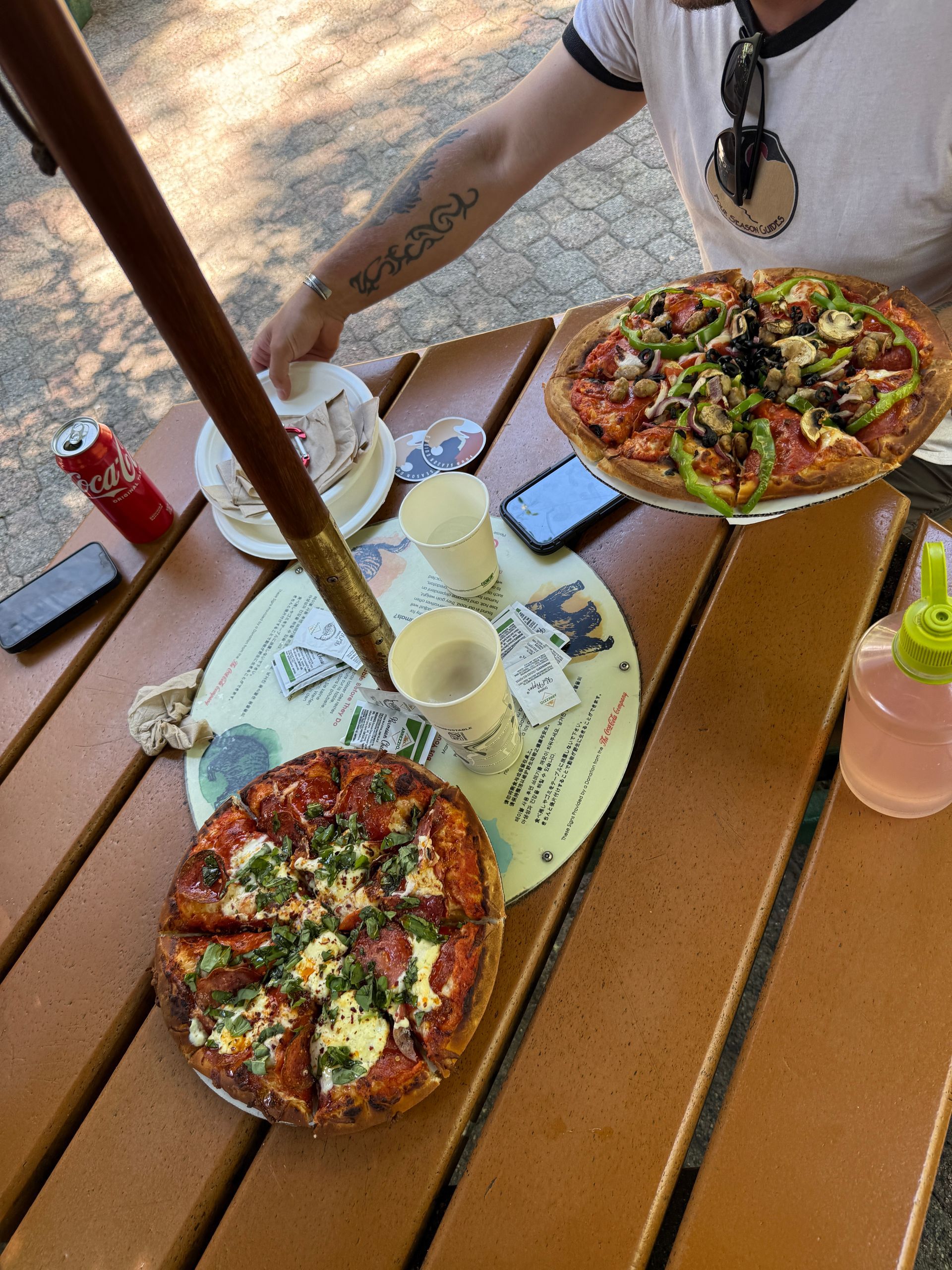 Two pizzas on a table with a person reaching for a small plate. Outdoor setting.