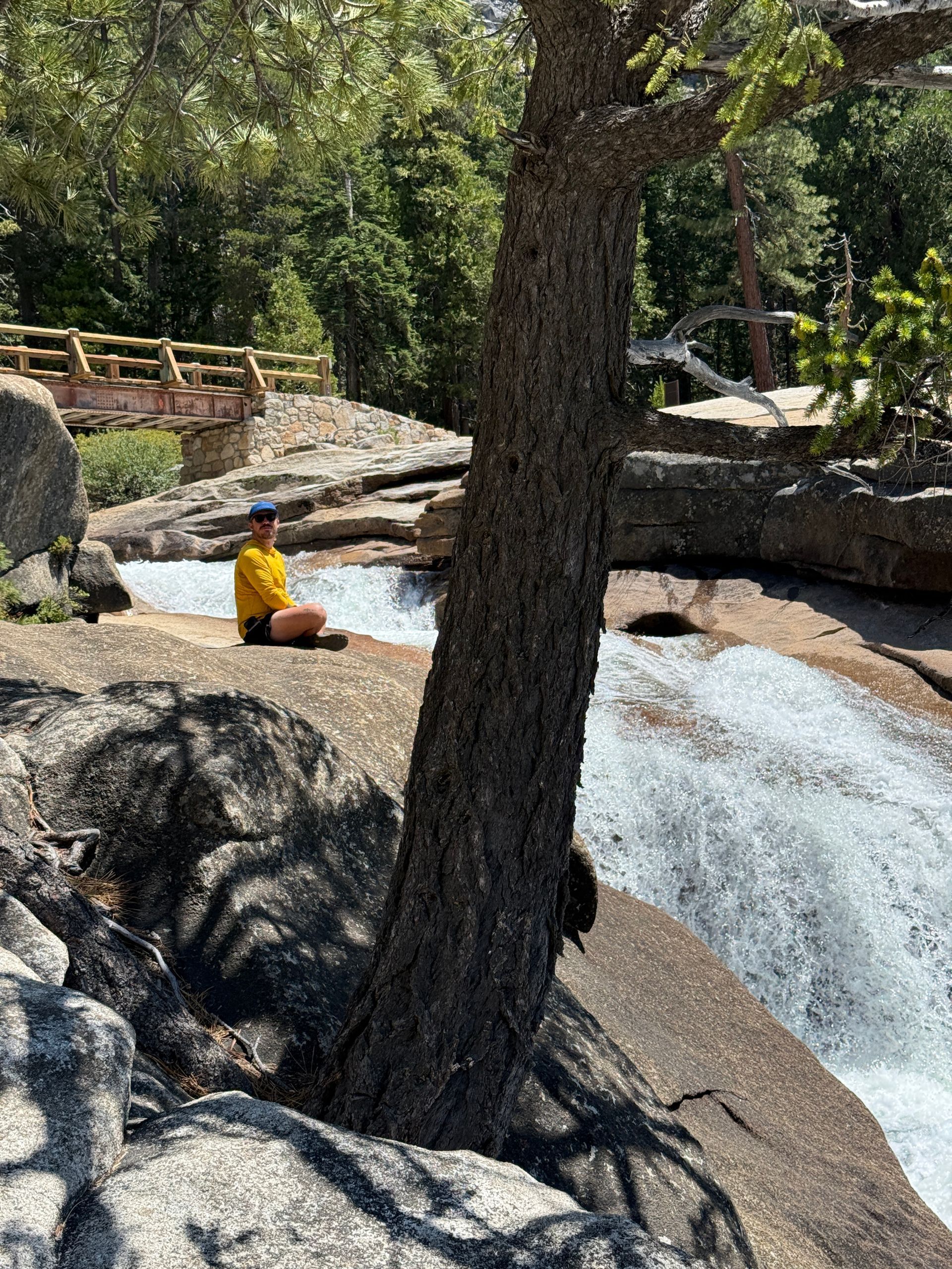 Person sitting in meditation pose near waterfall, tree in foreground. Yellow shirt, sunny day.