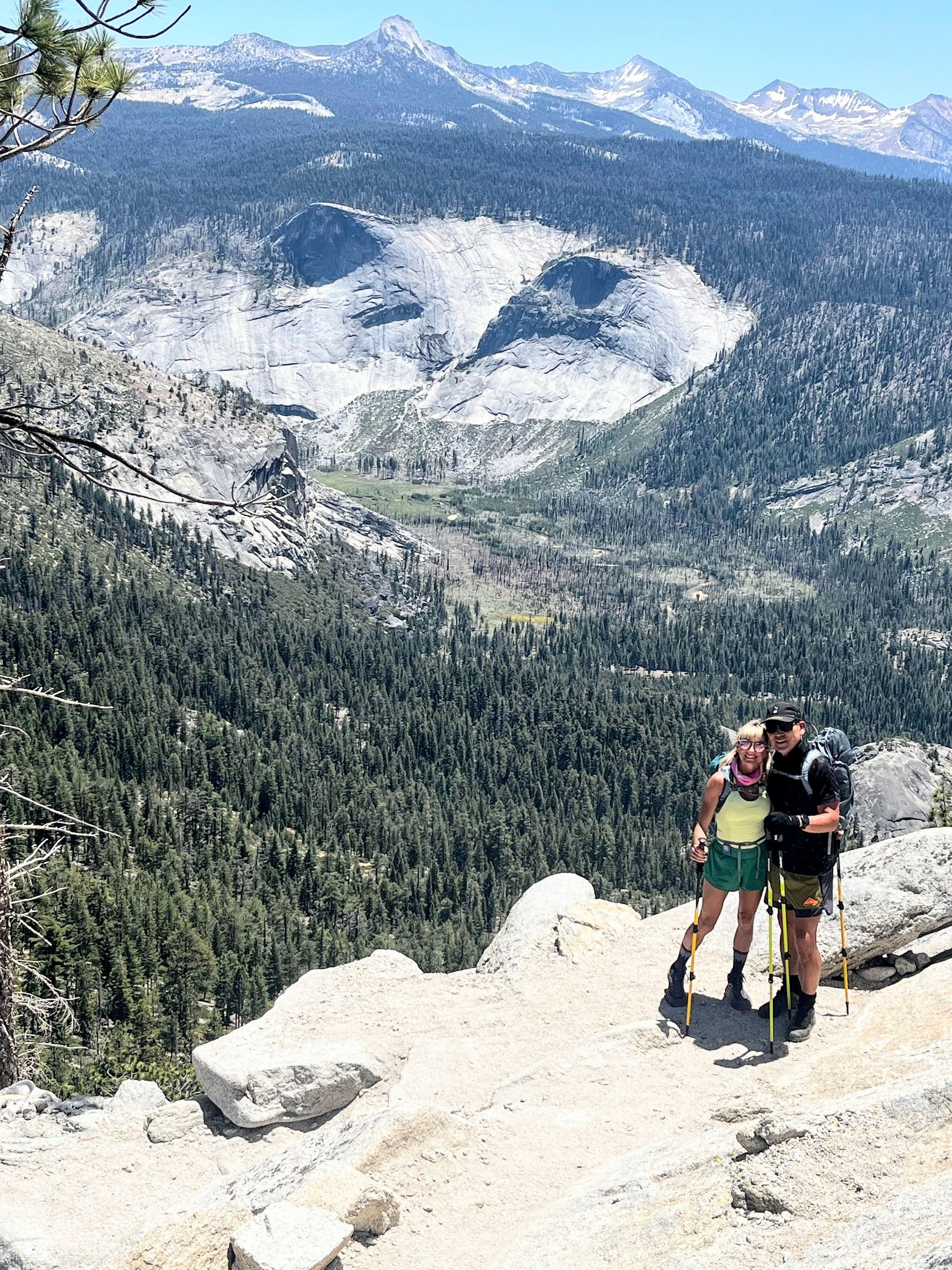 Couple stands on a cliff, overlooking a valley. They are hiking in the mountains. Sunny day.