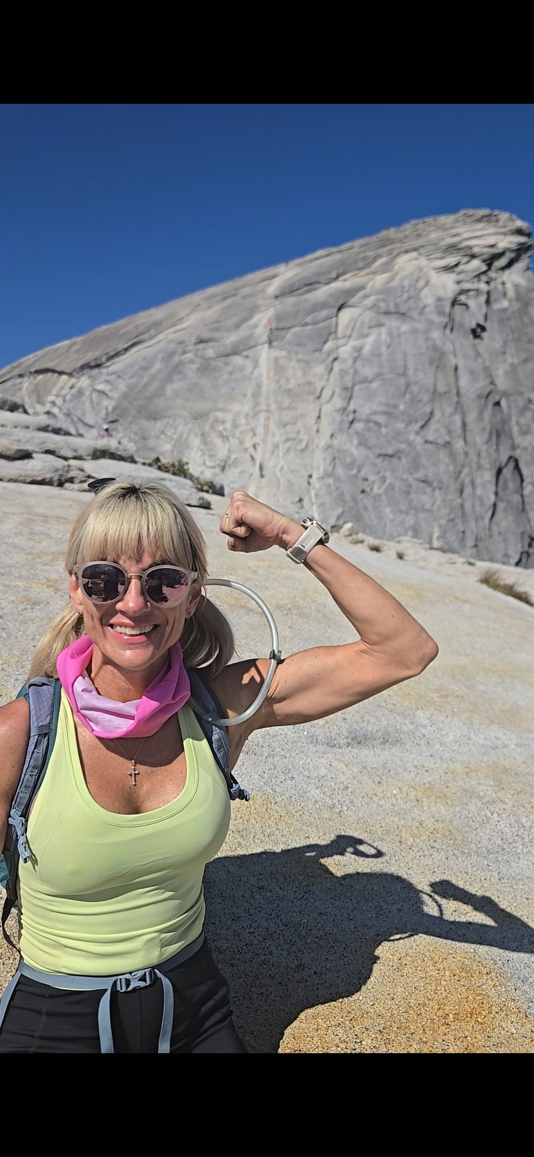 Woman flexing her bicep in front of a mountain. Wearing sunglasses, pink neck gaiter, and backpack.