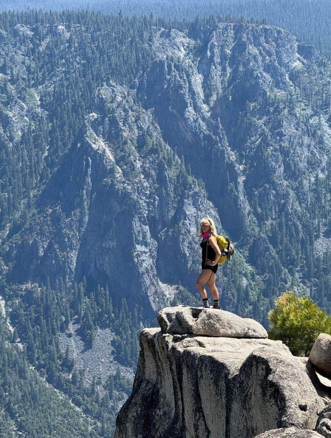 Woman stands on a rocky cliff, overlooking a forest-covered mountain range.