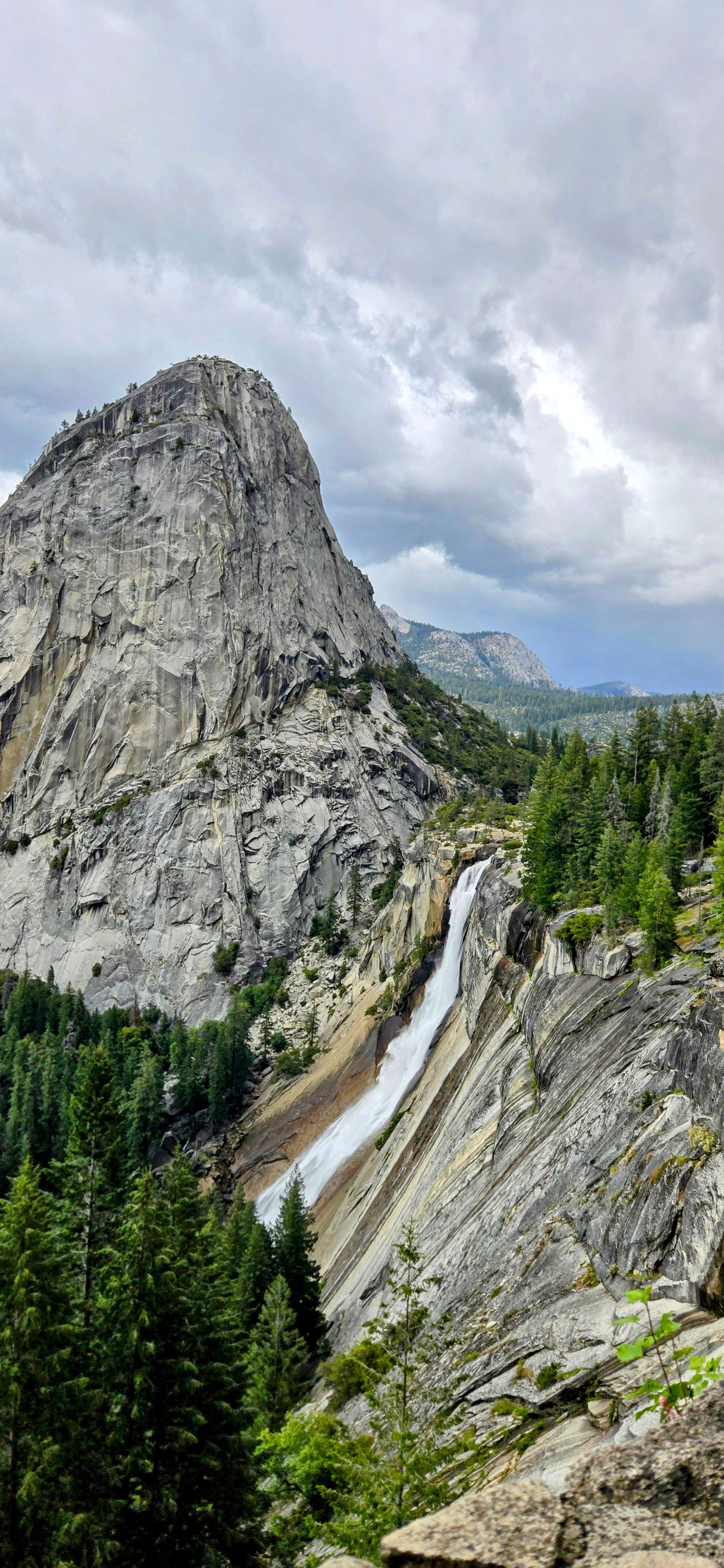 A waterfall cascades down a rocky cliff face, with a large mountain peak in the background. Green trees dot the landscape.