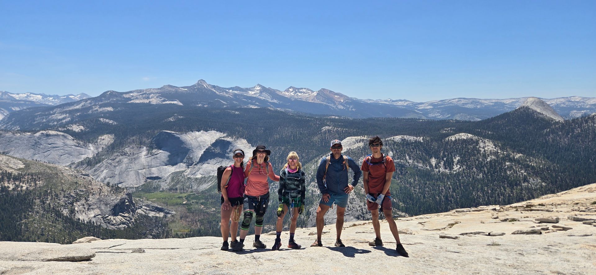 Five people stand on a rocky mountain, posing for a photo. A mountainous landscape stretches behind them under a bright blue sky.