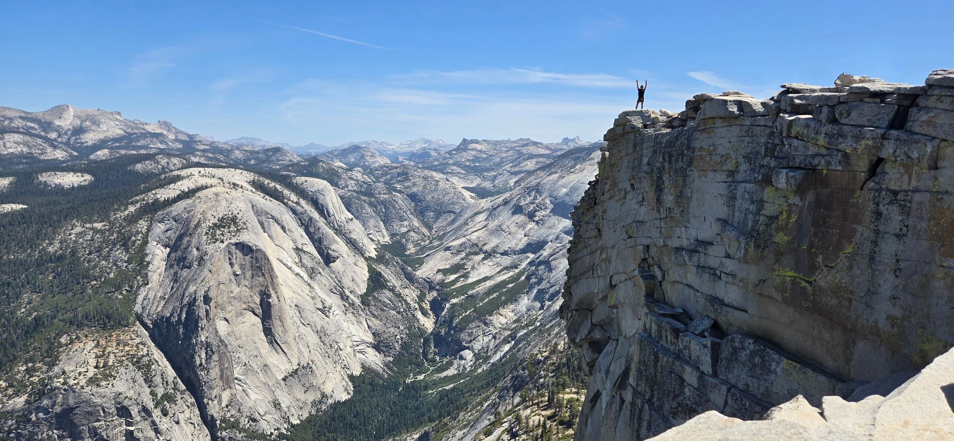 Person stands on cliff edge, arms raised, overlooking mountain range with clear blue sky.