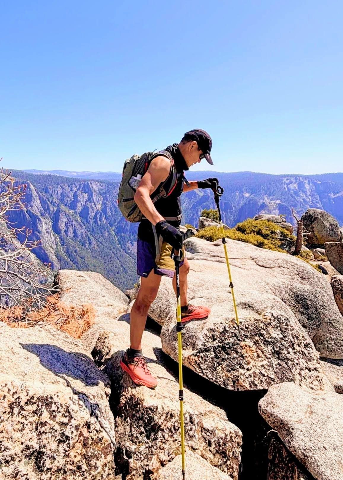 Hiker on rocky summit, using trekking poles. Backpack, shorts, and cap. Mountain vista backdrop under blue sky.