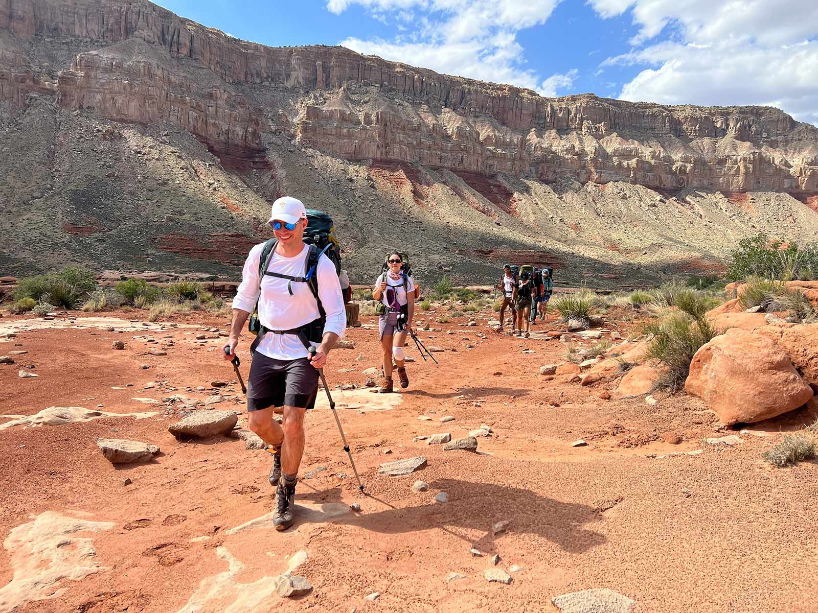 Hikers on a red trail in a canyon, under a sunny sky. Man in front, others following.