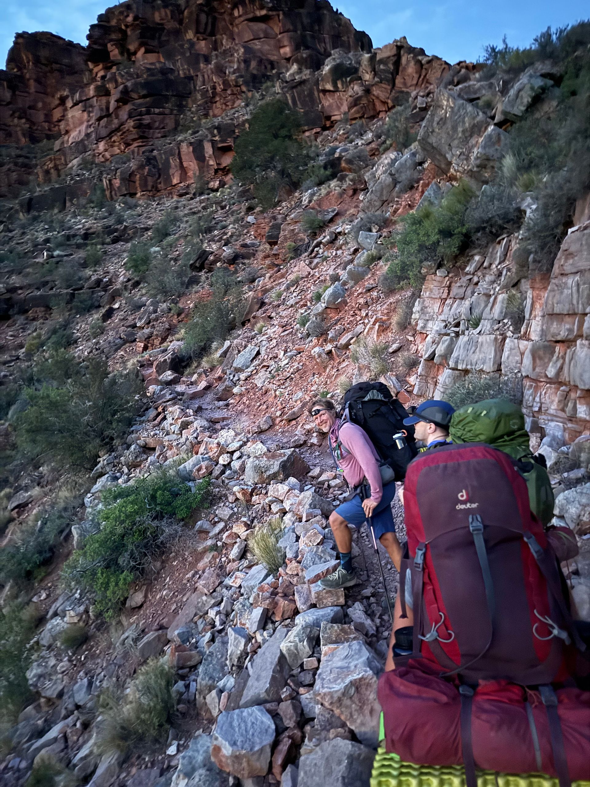 Two people hiking up a rocky mountain trail, carrying backpacks; reddish-brown cliffs in the background.