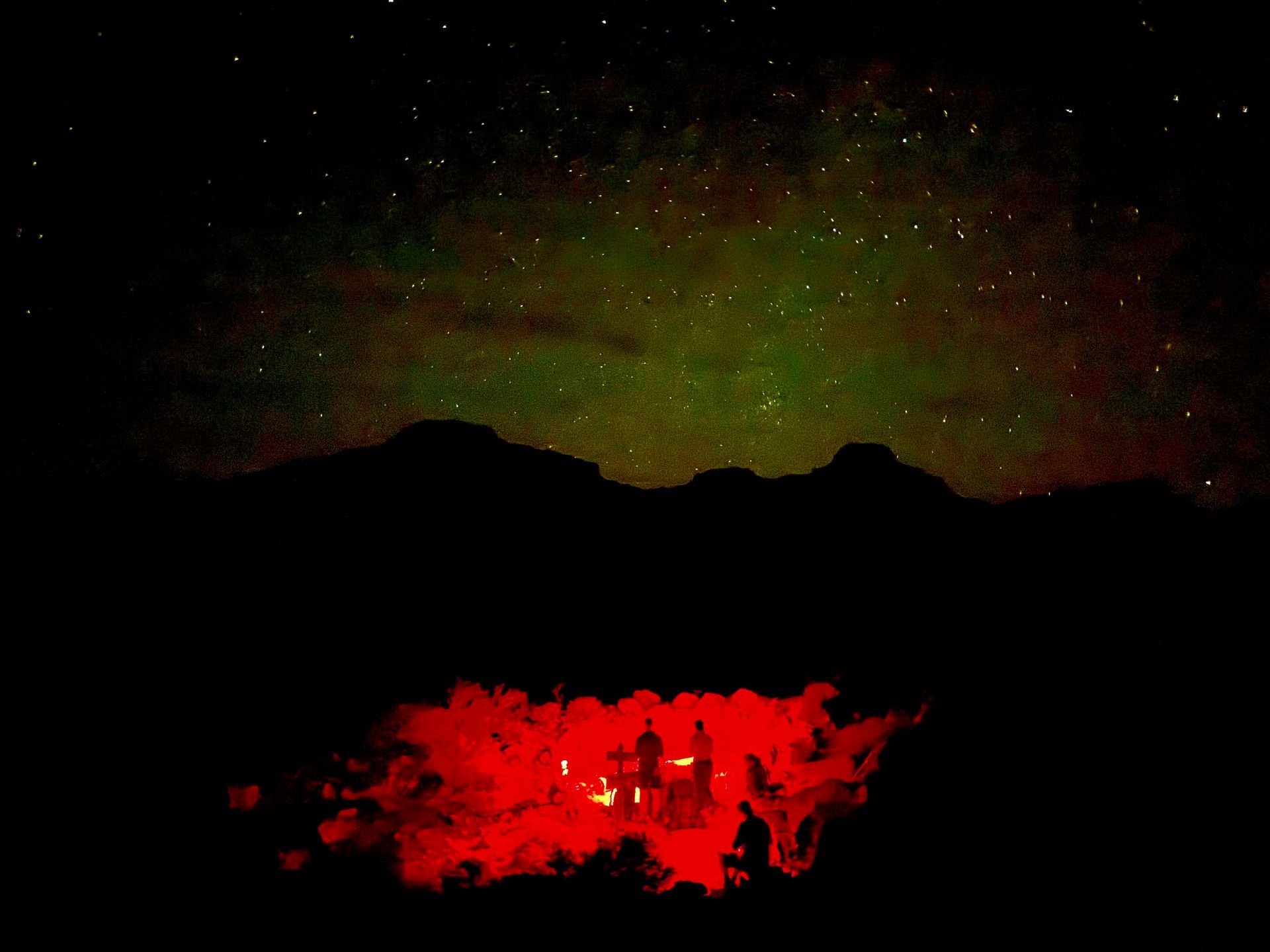 Night scene: Stars above green and black mountains. Red-lit structure reflects in water below.