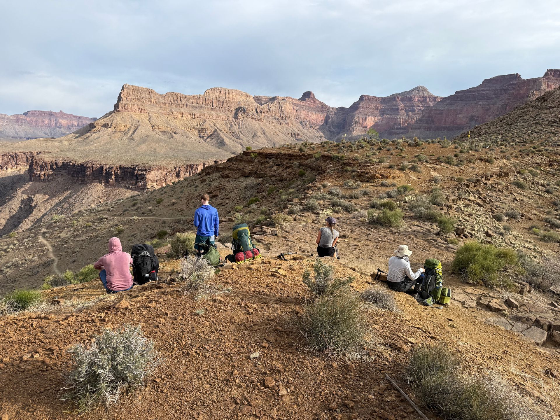 Hikers with backpacks on a brown, rocky trail overlook a canyon. Mountains fill the background under a cloudy sky.