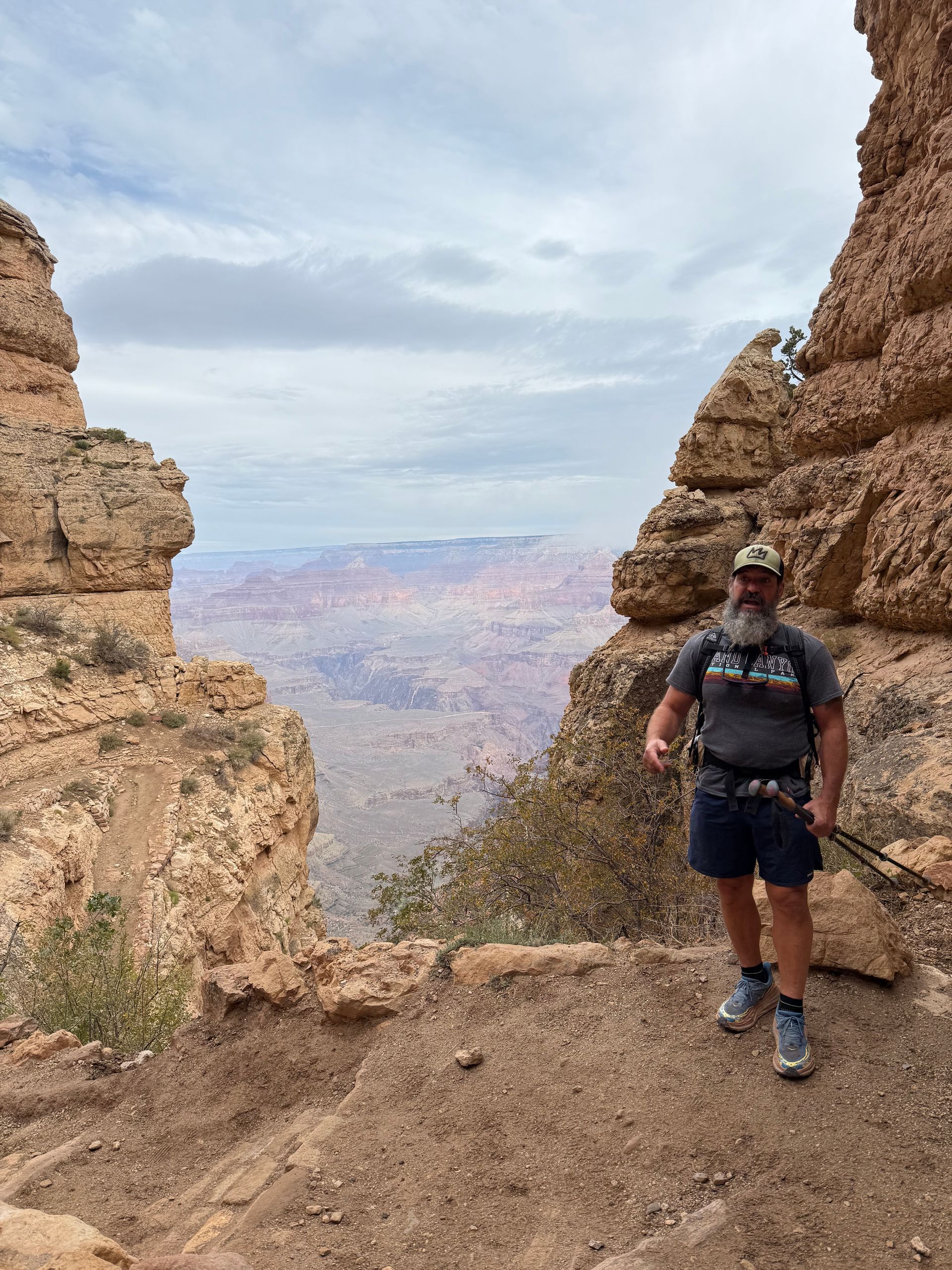 Man standing on a canyon edge, looking at a vast landscape. Cloudy sky, rock formations, and arid terrain.