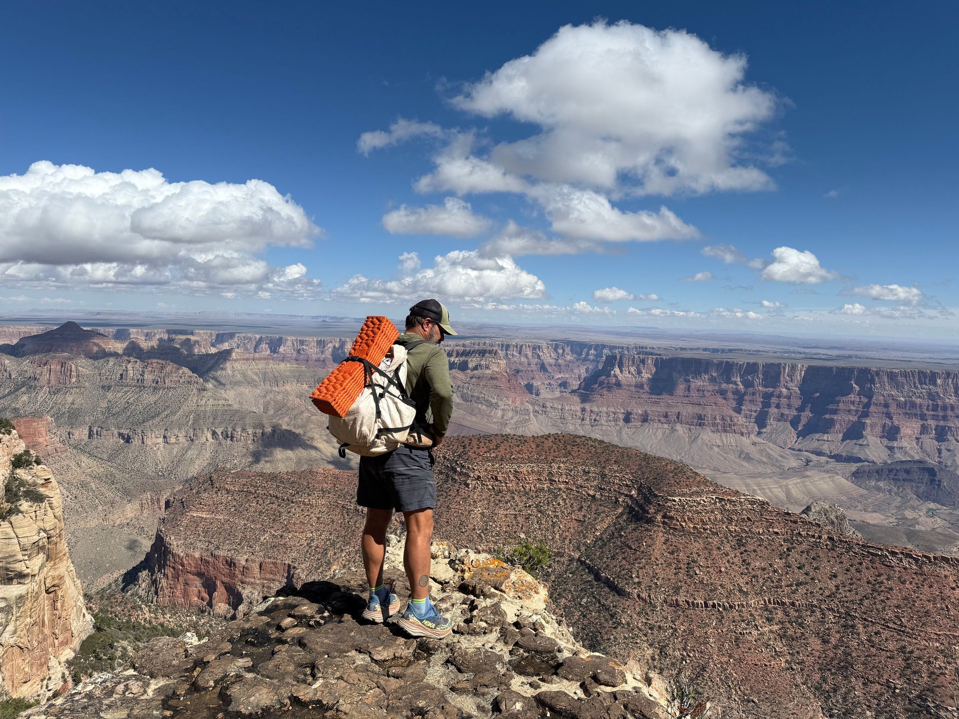 Hiker stands on a cliff overlooking the Grand Canyon; blue sky, white clouds, tan rock formations.