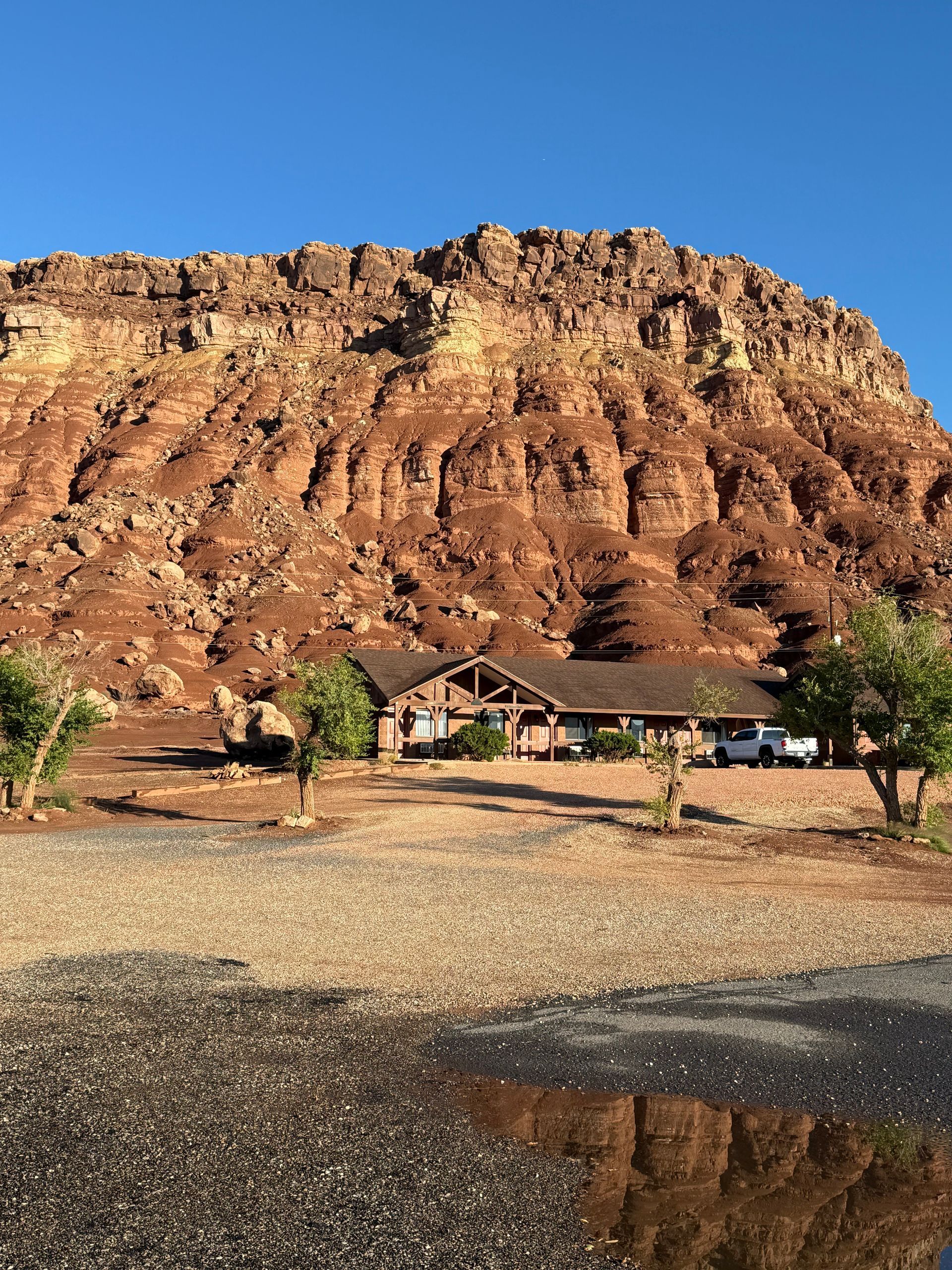 A cabin sits in front of a red rock mountain under a clear blue sky, gravel lot in foreground.