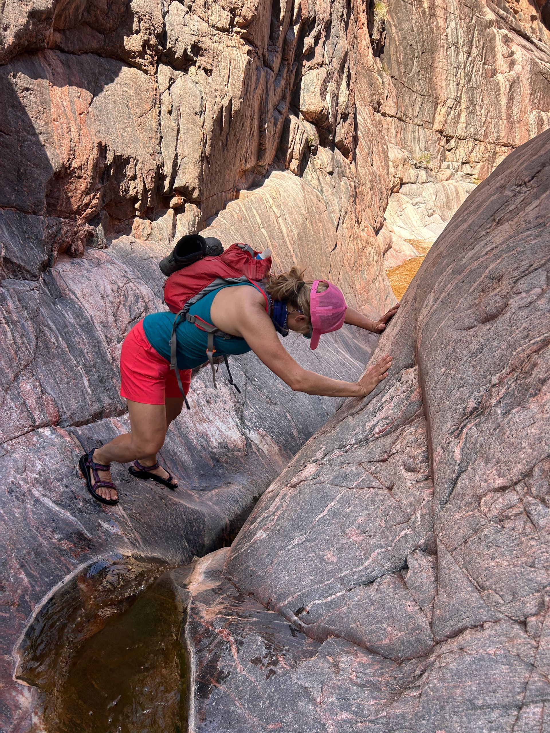 Person climbs a steep rock face in a canyon, wearing a backpack, red shorts, and a pink hat.