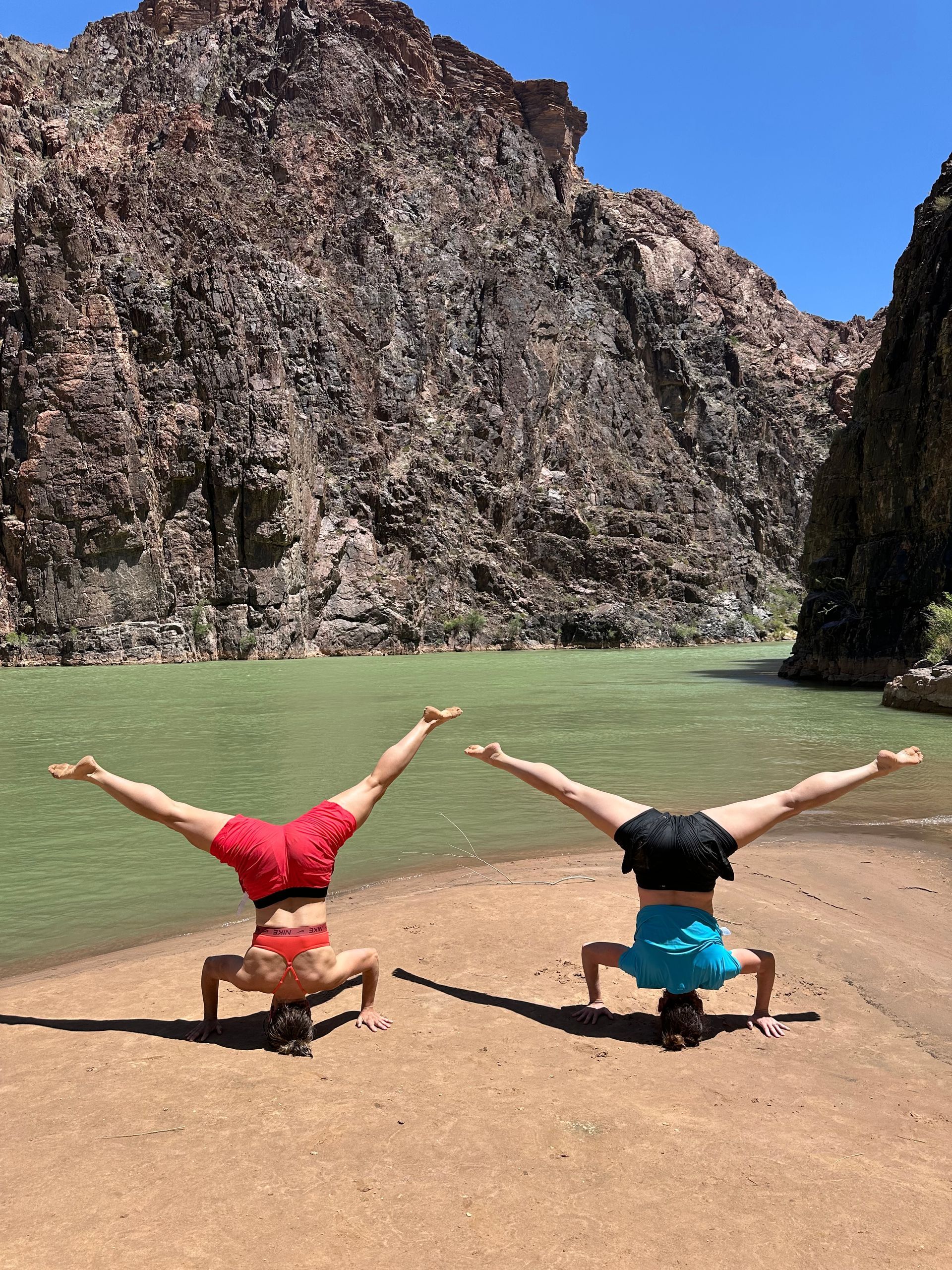 Two people doing headstands on a beach by a green river, with a rocky canyon in the background.