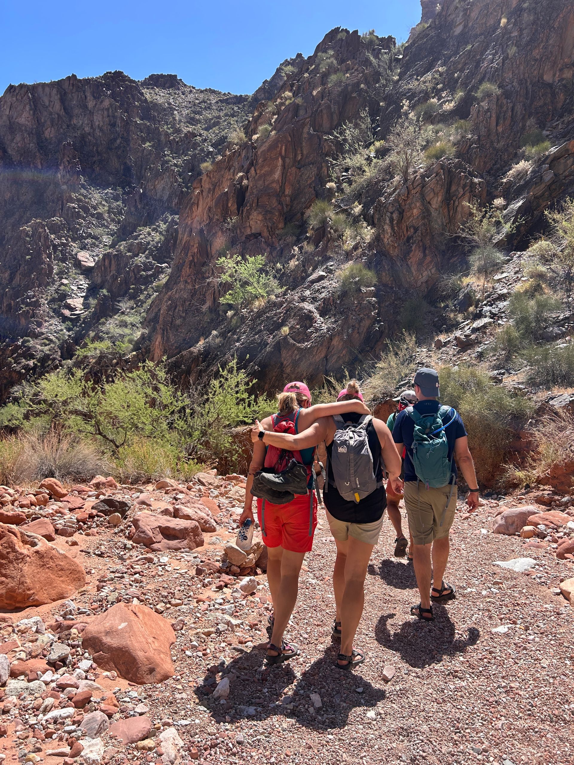 Three people hike on a red-rock trail through a canyon. Two have their arms around each other. Sunny day.