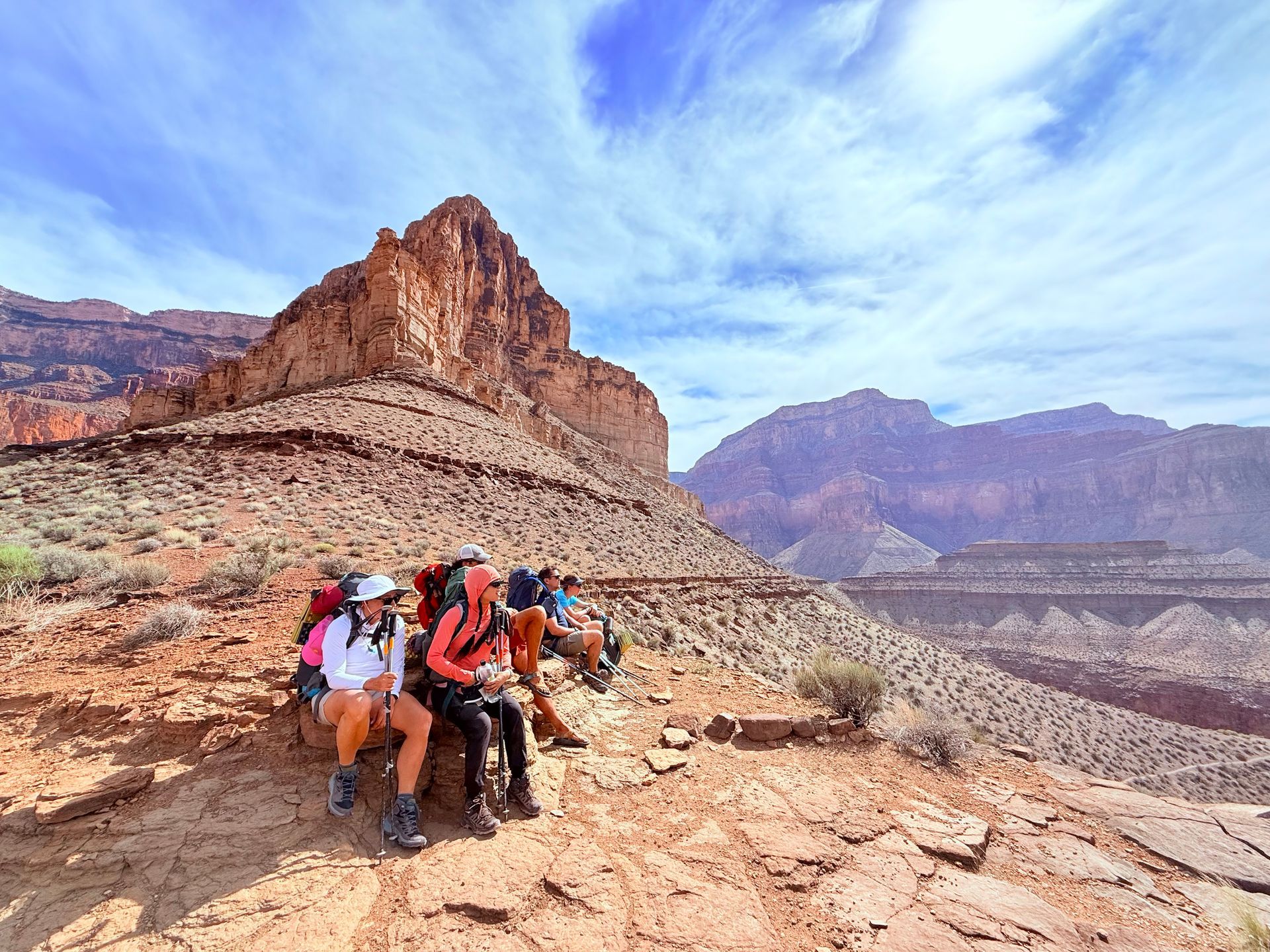 Hikers with backpacks resting on a ridge overlooking a canyon; red rock, blue sky.
