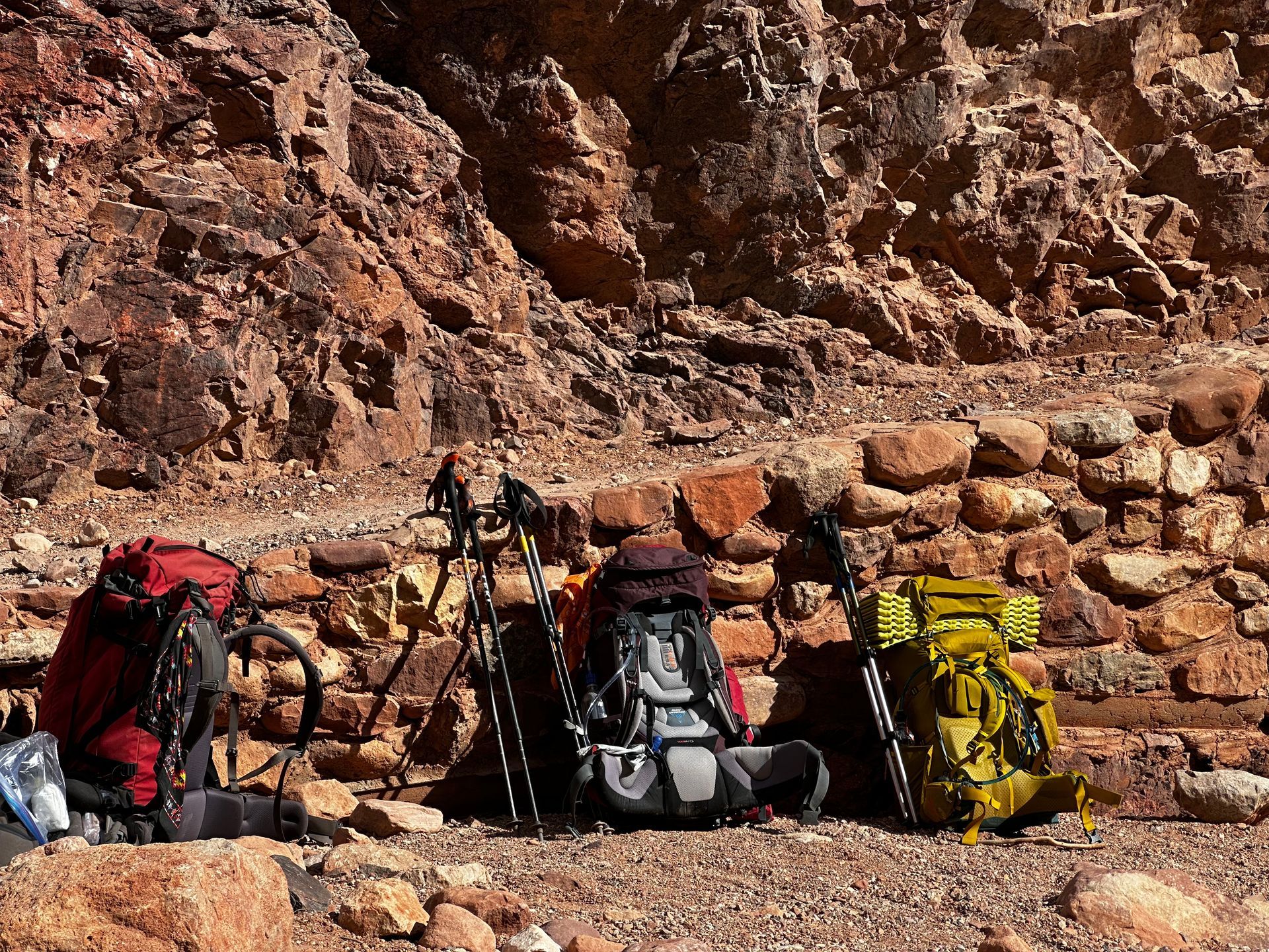 Three backpacks are sitting on the ground in front of a rock wall.