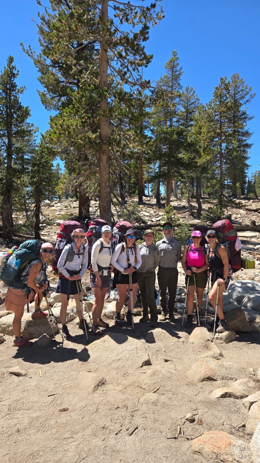 Group of hikers with backpacks and hiking poles pose with a park ranger on a sunny mountain trail.