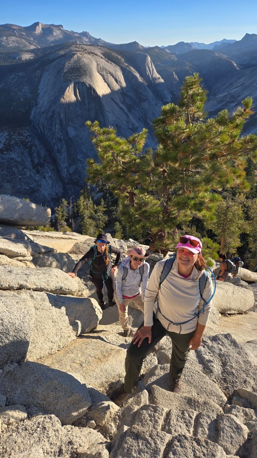 Hikers ascending a rocky trail with Half Dome in the background. Sunny day, pine trees, backpacks.