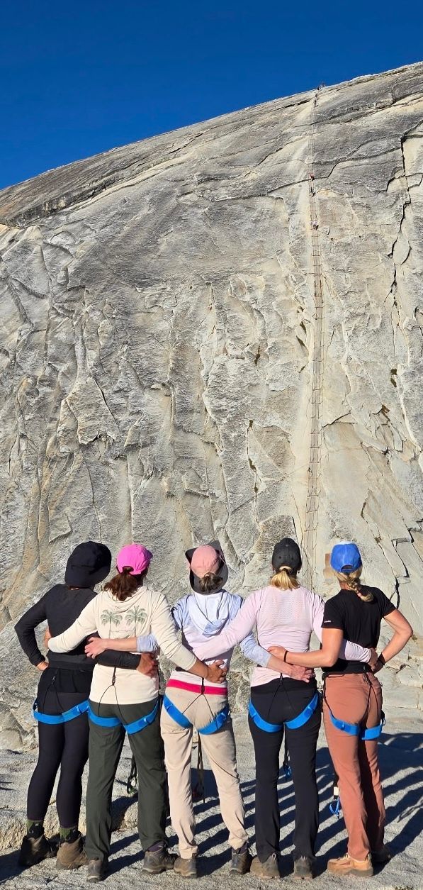 Five people with climbing harnesses stand embracing, facing a rock face. Blue sky overhead.