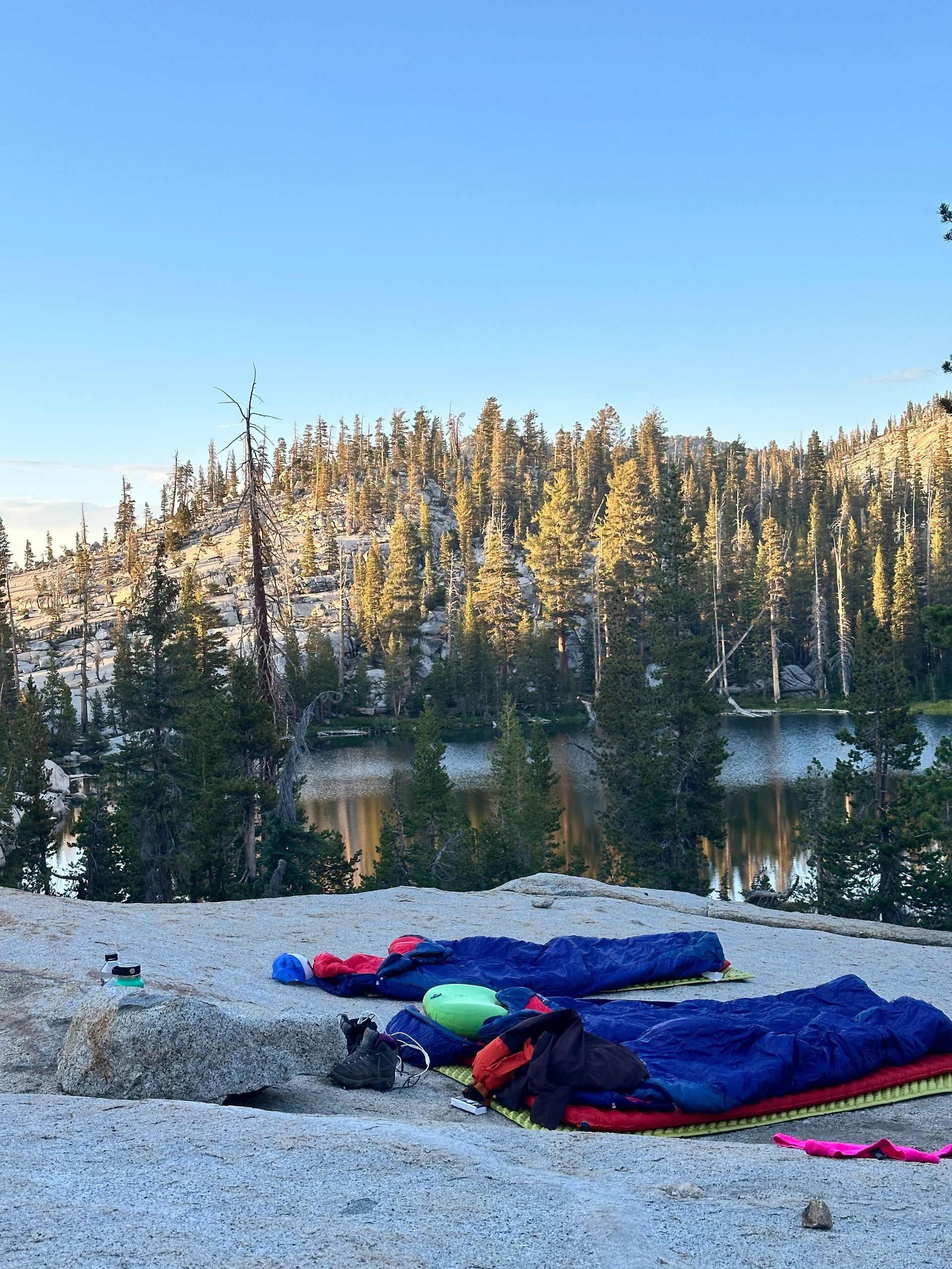Camping scene: sleeping bags on a rocky outcropping overlooking a lake and forested hills.