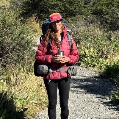 A woman with a backpack is standing on a dirt path holding a cup of coffee.