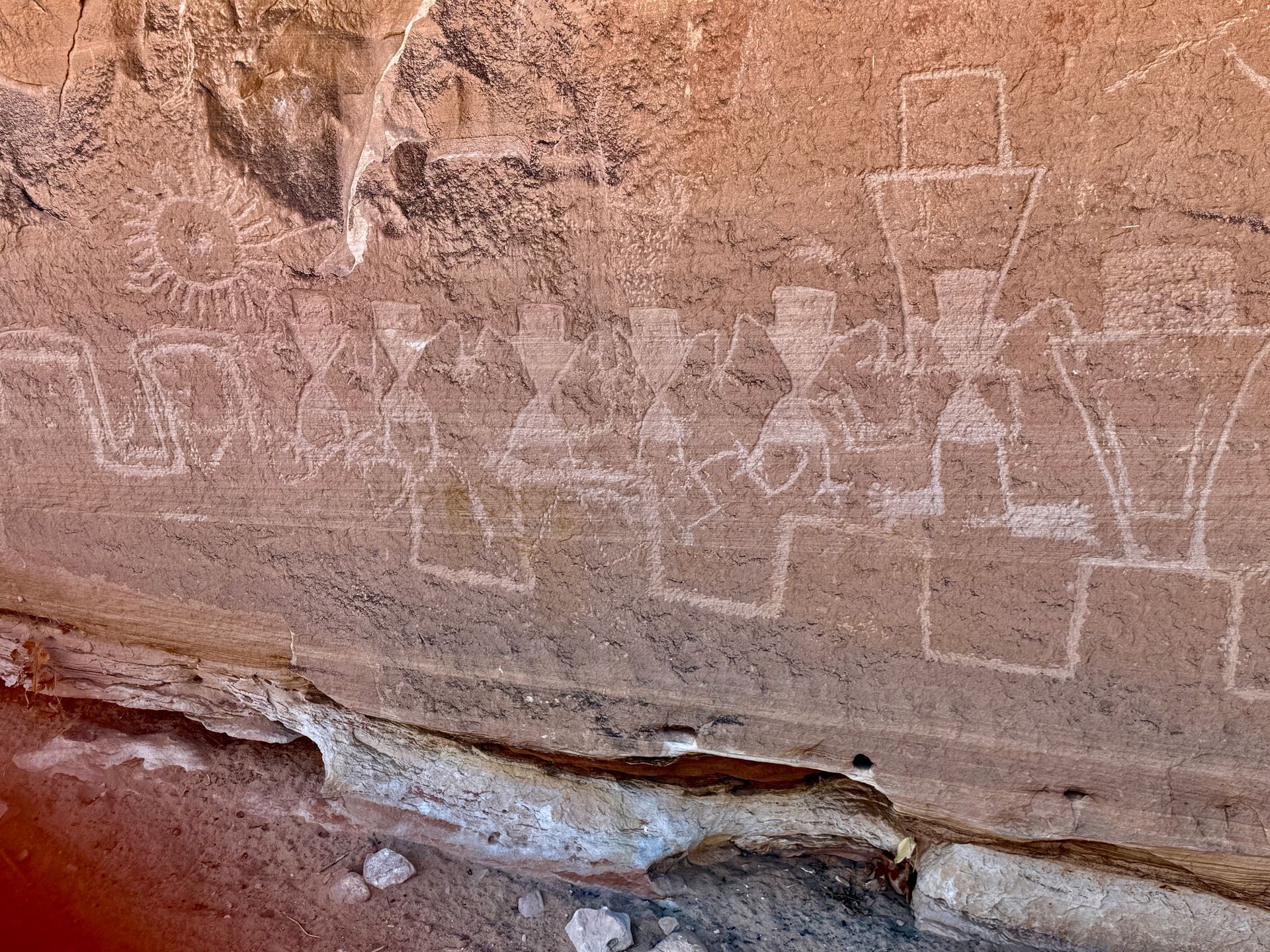 Rock art panel featuring human figures with geometric headdresses and a sun, etched into a sandstone surface.