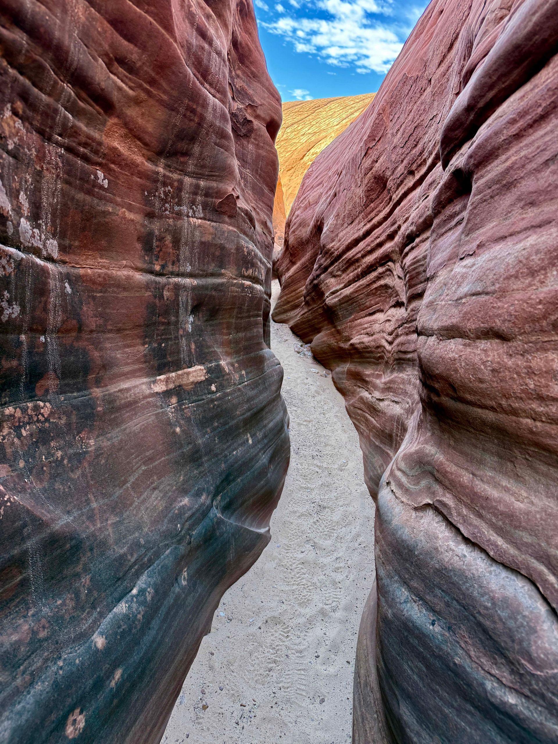 Narrow canyon with red and brown sandstone walls, sandy path, blue sky.